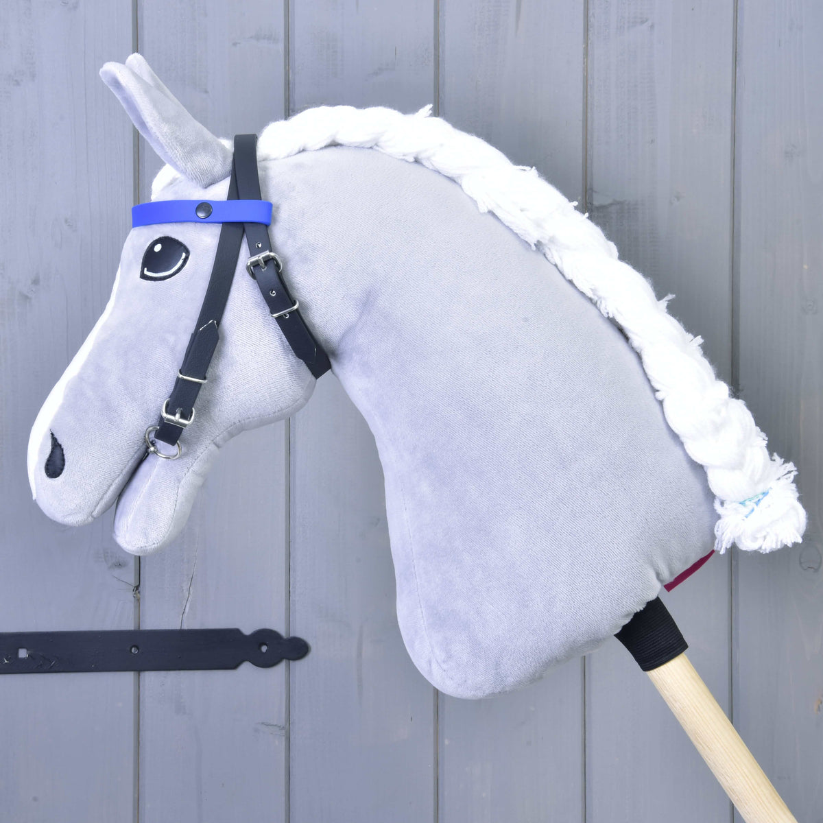 A blue "Bridle with 2 Headbands" (size M) by Hobby Horse LarDen is on a plush hobby horse with a white yarn mane, attached to a wooden stick and resting against a gray wooden wall.