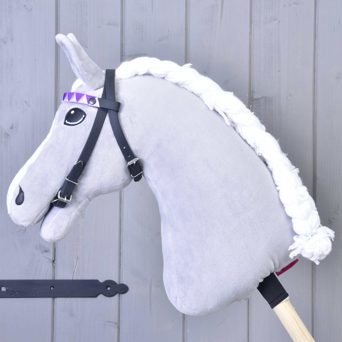 The Hobby Horse LarDen Bridle with 2 headbands purple (size M) features a gray plush horse with a white braided mane, black bridle without noseband, and a purple diamond-patterned ribbon on a wooden stick, set by a light gray wooden wall.