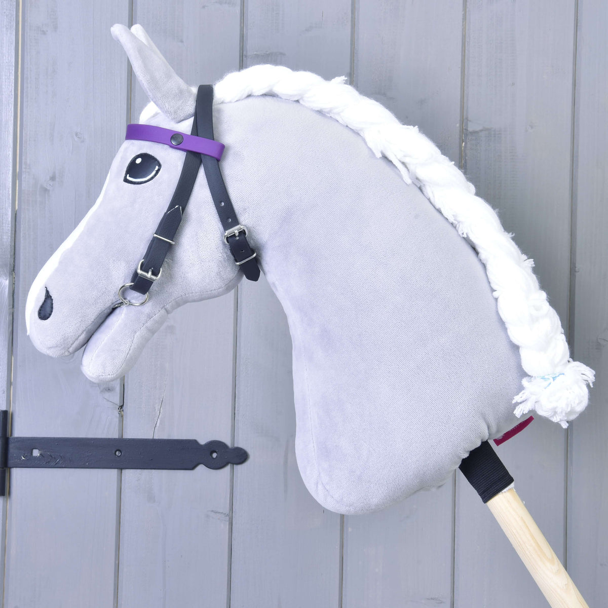 A gray hobby horse with a white yarn mane and a Hobby Horse LarDen bridle with 2 purple headbands (size M), without noseband, on a wooden stick, is positioned in front of a light gray wooden wall.