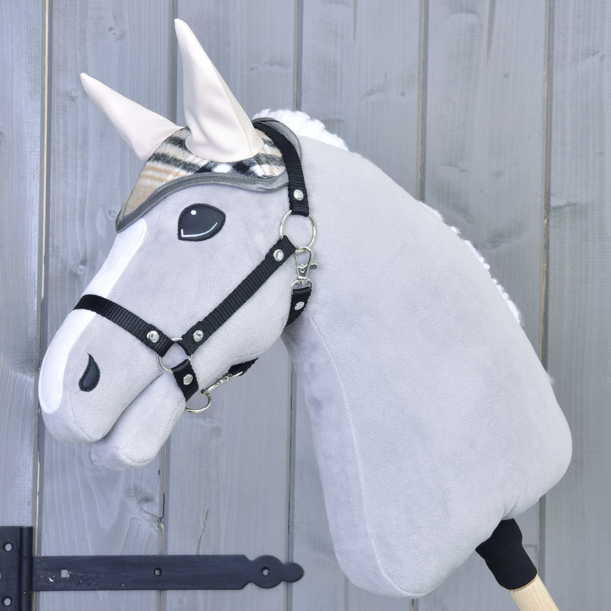 The Hobby Horse LarDen plush gray hobby horse wears the Ear bonnet Cremo England and a black halter, mounted on a wooden stick in front of a light gray wooden fence.