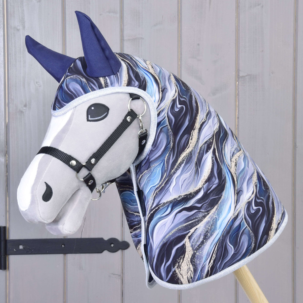 The Hobby Horse LarDen Raincoat Galaxy Cobalt features marbled blue, white, and black fabric, dark blue ears, a black halter, and matching accessories on a wooden stick, shown against a light wood wall.