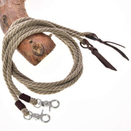 The Split Western Reins Dallas by Hobby Horse LarDen is a beige, coiled braided rope with leather accents and two metal snap hooks at the ends, displayed draped over wood on a white background.