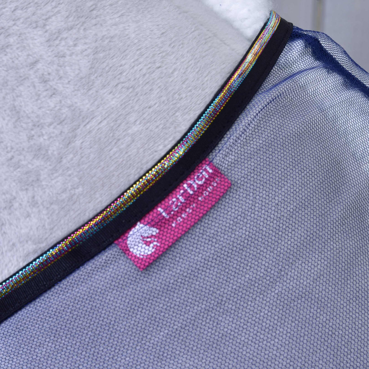 Close-up of Fly sheet LarDen by Hobby Horse LarDen, featuring PES mesh fabric, iridescent trim, and a pink HARPER tag with white logo. Soft white material in the background highlights its lightweight design for hobby horse accessories.