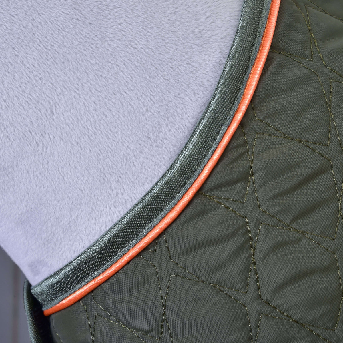 Close-up of the Blanket Sport by Hobby Horse LarDen, a quilted green protective horse blanket with orange piping and visible stitching, shown lying on a soft, light grey textured surface.