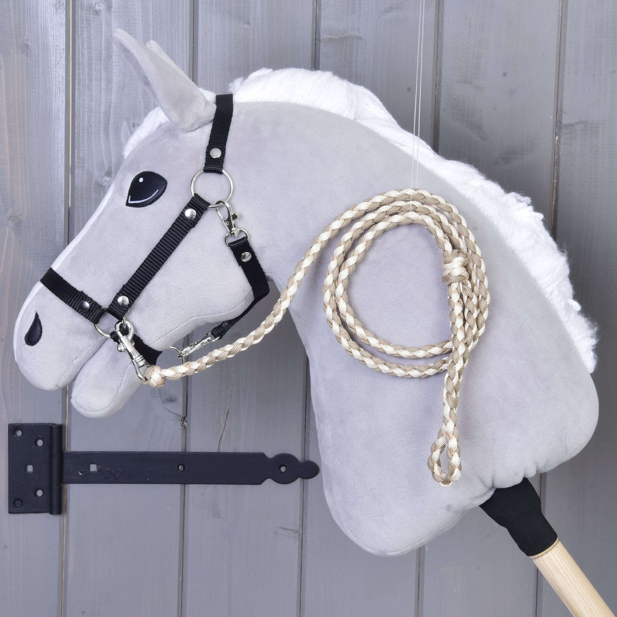 The Hobby Horse LarDen Hobby Horse – Knitted Lead Rope Double Latte, a gray stick horse with black bridle and braided reins, is displayed against a gray wooden wall.