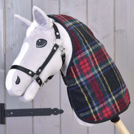 A plush hobby horse with a white face and black bridle wears the Hobby Horse LarDen Stable Blanket Edinburgh in red and green plaid, posed before a gray wooden wall with a visible black metal hinge.