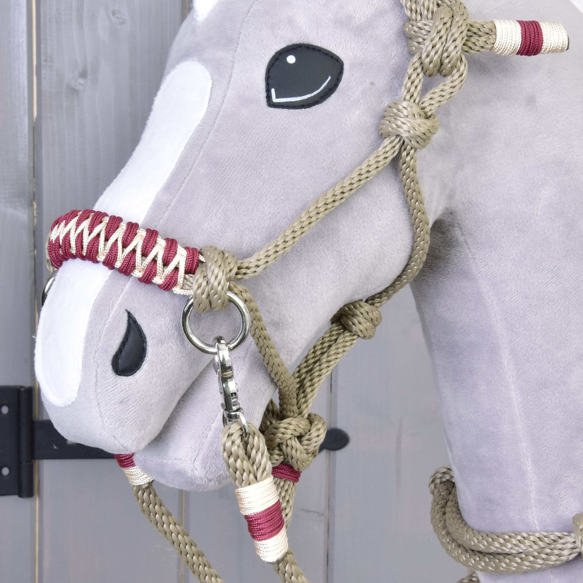 A close-up of the Hobby Horse LarDen’s head wearing the Rope set Eleganza—a soft, gray hobby horse with black button eyes, white felt patches, and a beige and red halter—set against a wooden background.