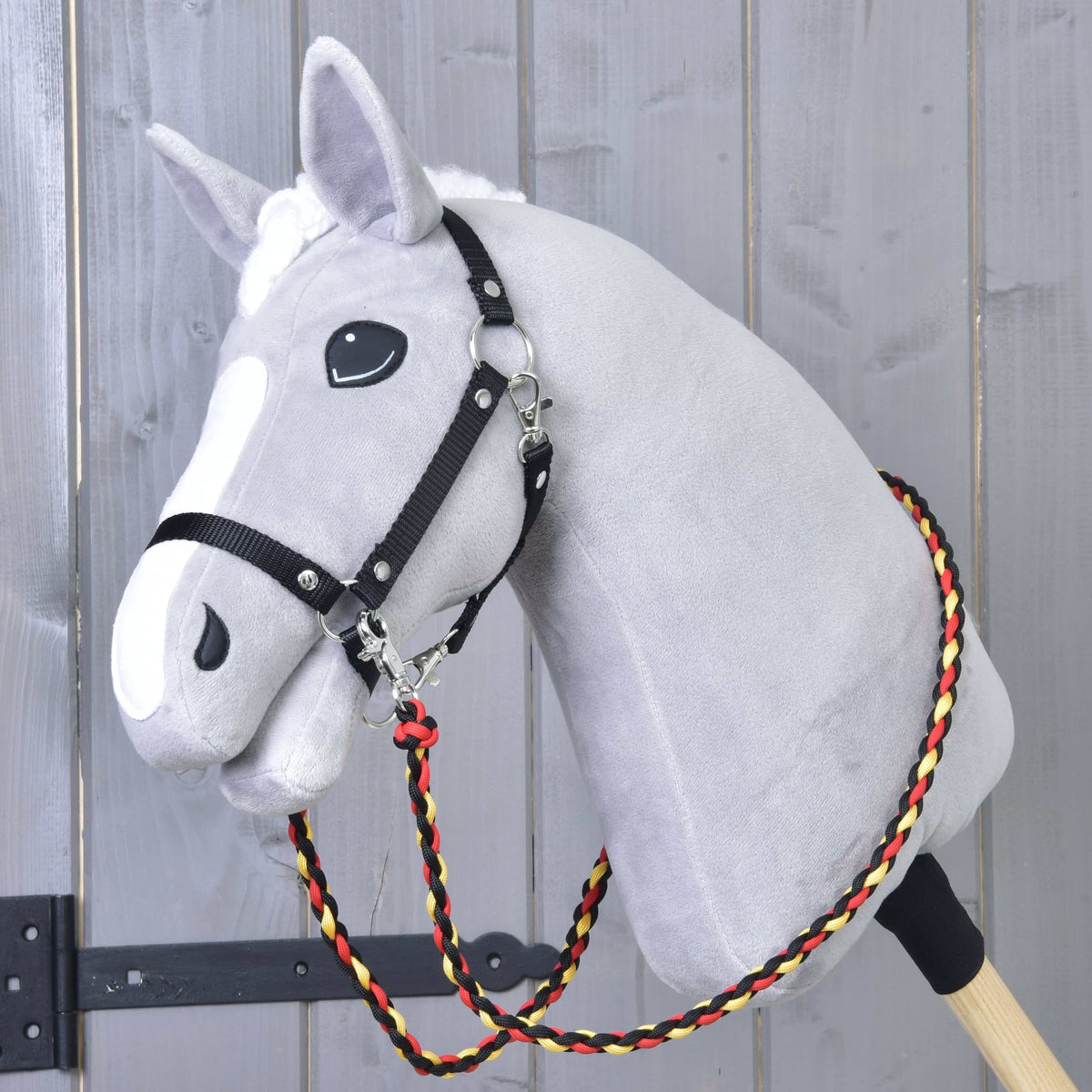 The Hobby Horse LarDen Reins Flag is a gray plush hobby horse on a wooden stick, with a black harness and handmade reins made of braided red, yellow, and black polypropylene rope, shown against a wooden wall.