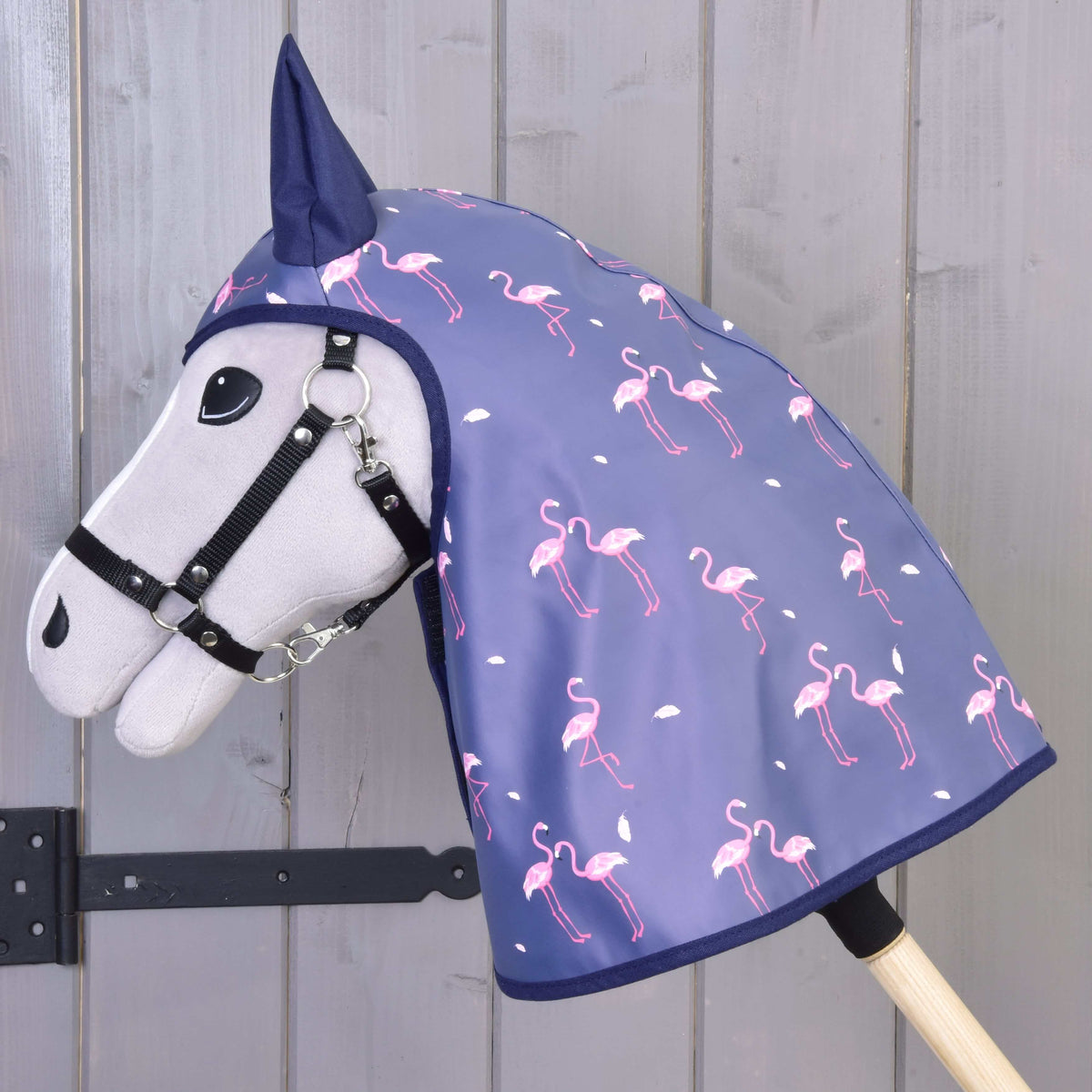 A gray Hobby Horse LarDen with a black bridle wears the Raincoat Flamingo—an all-weather blue hood with pink flamingos and white dots—set against a wooden background.