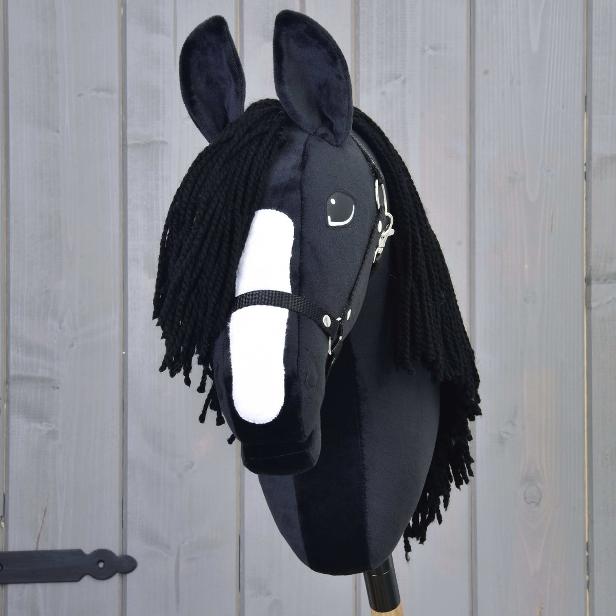 The Hobby Horse LarDen's Freya with Black Halter, inspired by the Irish Cob and featuring a long black yarn mane, stands before a gray wooden wall.