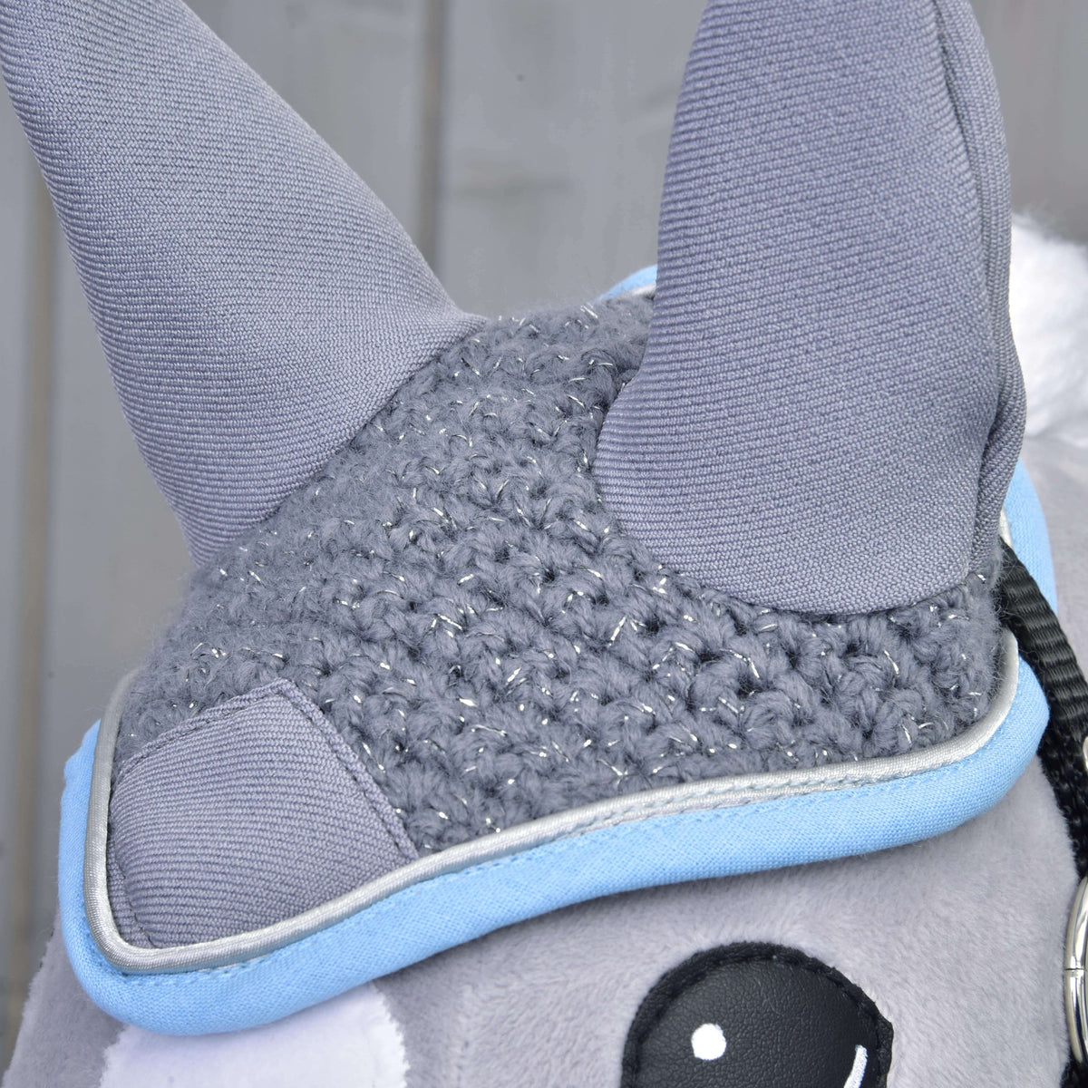 Close-up of a Hobby Horse LarDen plush toy wearing the “Ear bonnet crocheted grey,” featuring two gray crochet-style ears, a knitted gray patch with silver threads, light blue trim, and part of a black and white eye. Blurred background.
