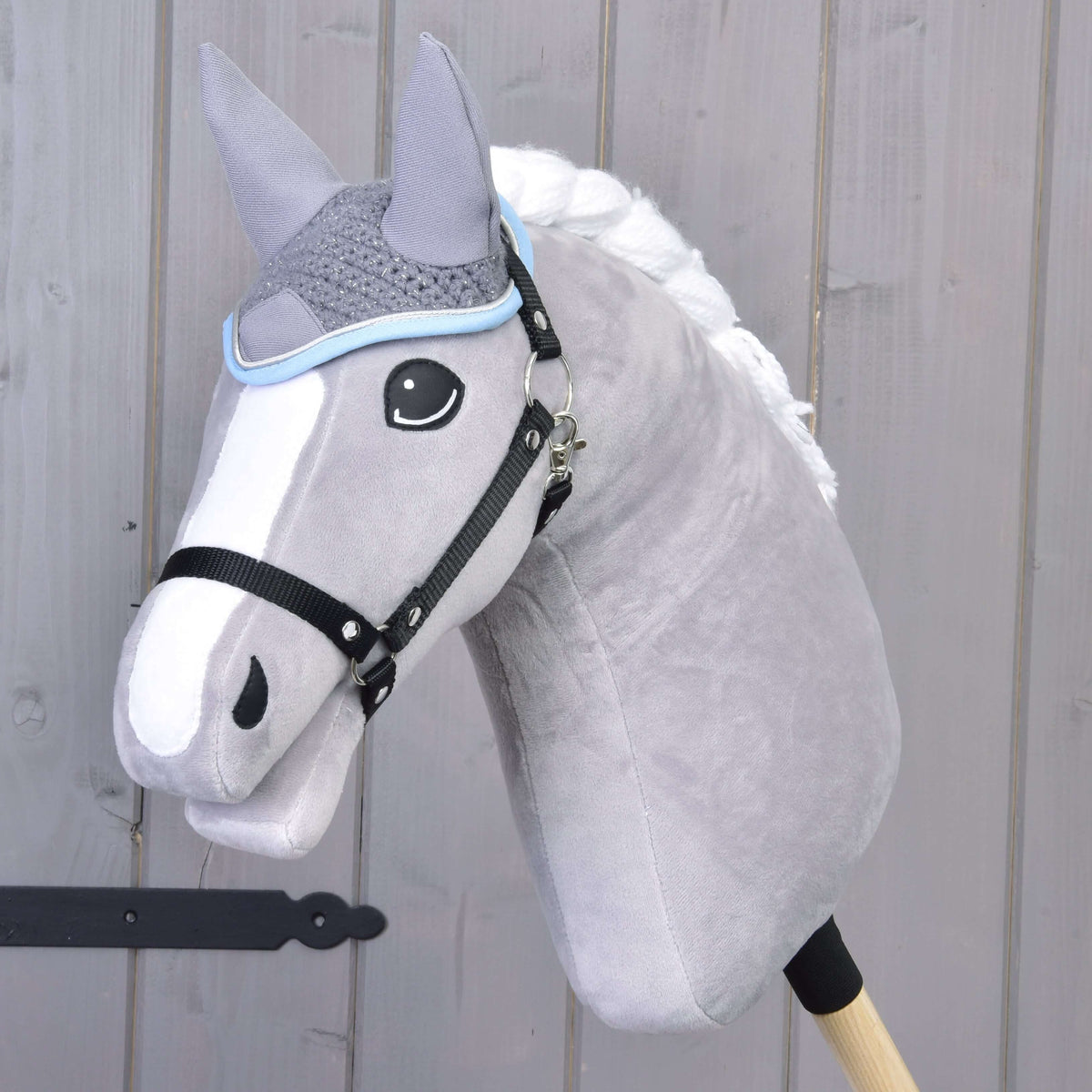 The Hobby Horse LarDen Ear Bonnet Crocheted Grey is showcased on a plush grey hobby horse with a white mane and black bridle, mounted on a wooden stick in front of a light wooden wall.