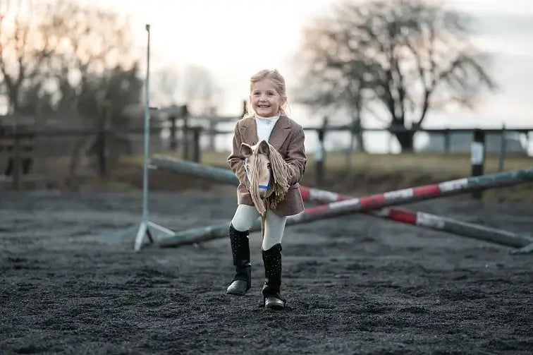 En smilende pige rider på sin kæphest på en ridebane udendørs med krydsede springbomme og træer i baggrunden. Hun har brun jakke, hvide bukser og sorte ridestøvler på.