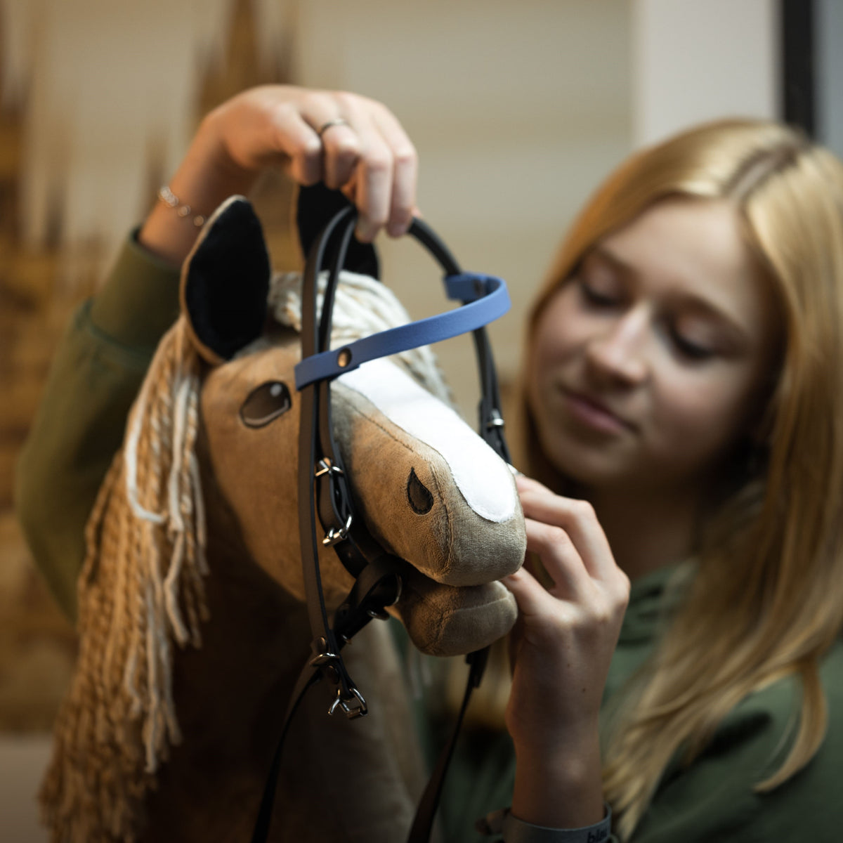 En pige med lyst hår sætter trense på sin Hobby Horse og justerer omhyggeligt remmene på kæphesten.