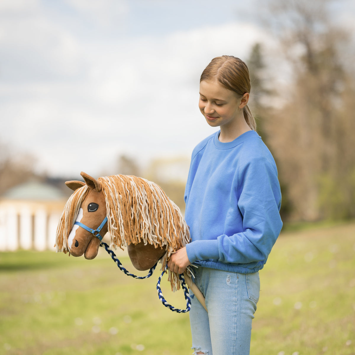 En ung person i blå sweatshirt og jeans står udenfor på en græsplæne og smiler ned til en brun kæphest med trense af reb.