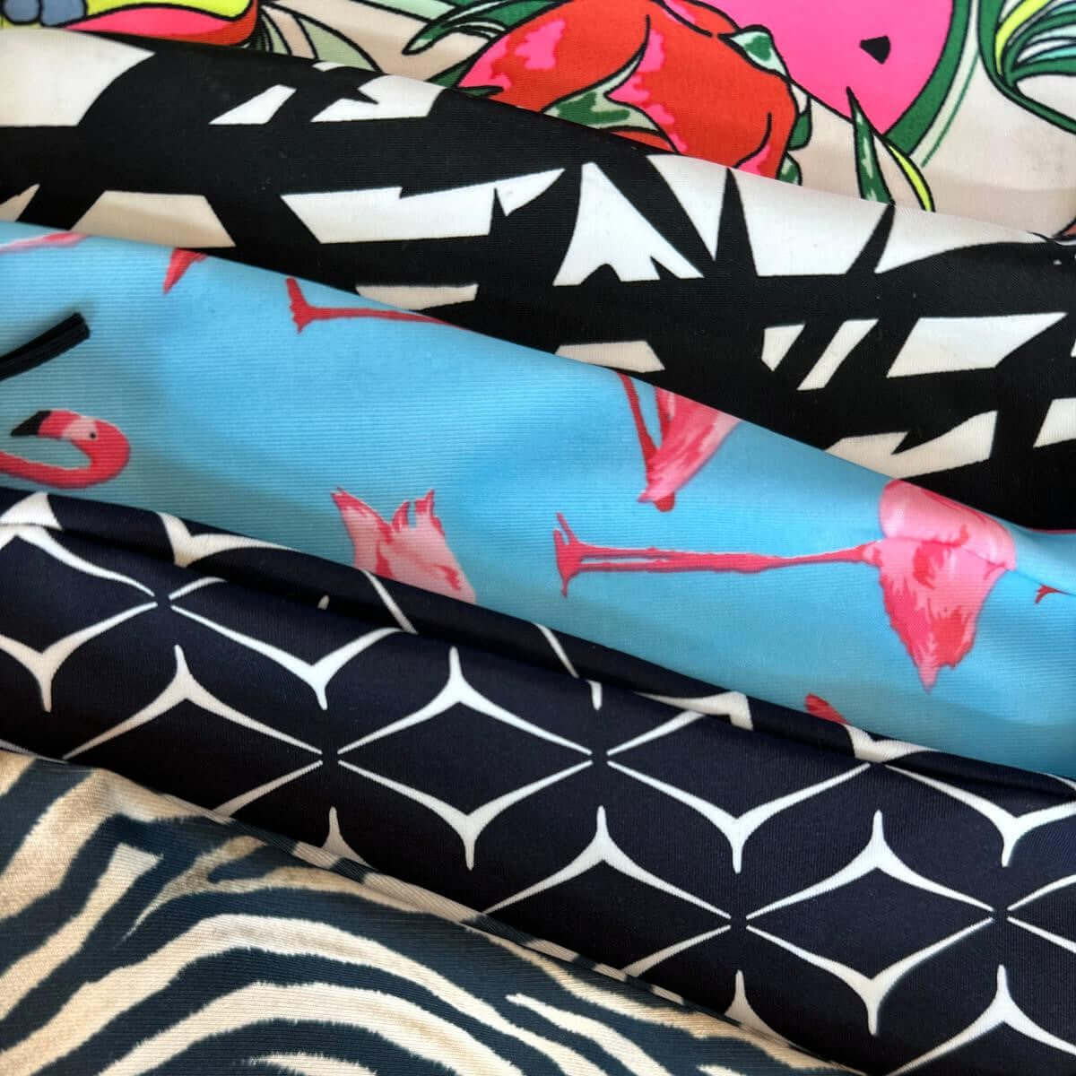 Fem stofruller med forskellige mønstre: farverig frugt, sort-hvid abstrakt tekst, blå med lyserøde flamingoer, marineblå med hvide geometriske figurer og zebrastriber.