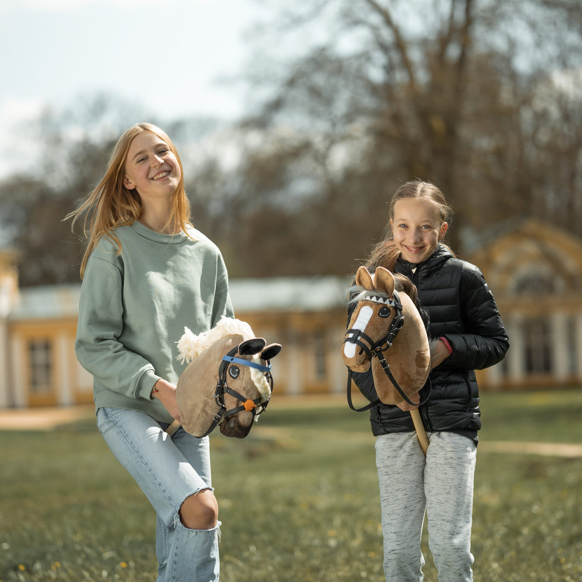 To smilende piger står udenfor med deres kæpheste, den ene i lysegrøn sweater og hullede jeans, den anden i sort jakke og grå bukser.
