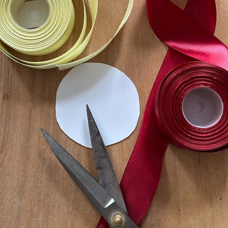 A pair of scissors, a circle of white paper, a roll of yellow ribbon, and a roll of red satin ribbon are arranged on a wooden surface, ready for your next Hobby Horse crafting session.