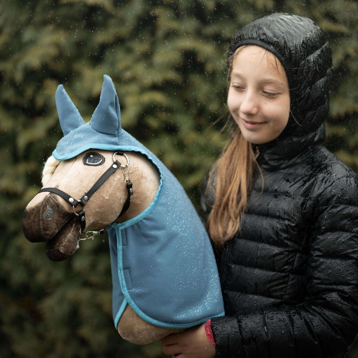 A smiling girl in a black hooded jacket stands outdoors in the rain, holding her cherished hobby horse plush toy dressed in blue, with green foliage blurred in the background—a perfect moment for any hobby horsing enthusiast.