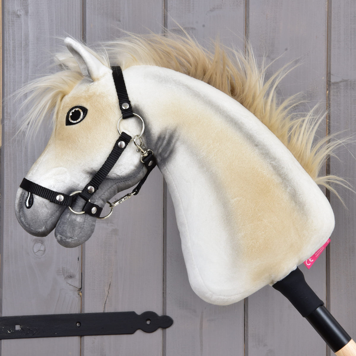 The Hobby Horse LarDen "Hobby Horse Honey Pearl with Black Halter" (size S) is a plush stick horse in beige and gray with a fluffy mane, black halter, and mounted on a stick, displayed before a wooden wall.