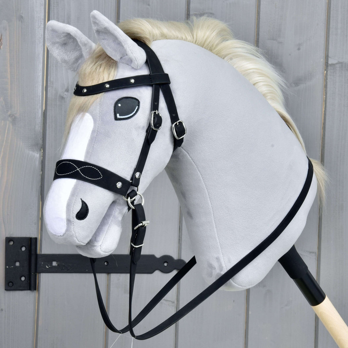 The Hobby Horse LarDen Complete bitless bridle, decorated, is shown on a gray plush hobby horse with a white mane and detachable reins, mounted on a wooden stick against a gray wooden background.