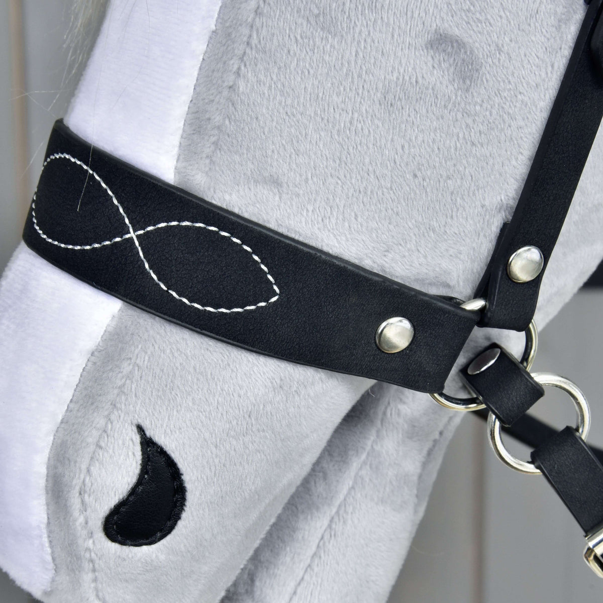 Close-up of a grey plush hobby horse’s head wearing the Hobby Horse LarDen Complete bitless bridle, decorated with white infinity-symbol stitching and a teardrop-shaped black patch near its eye.