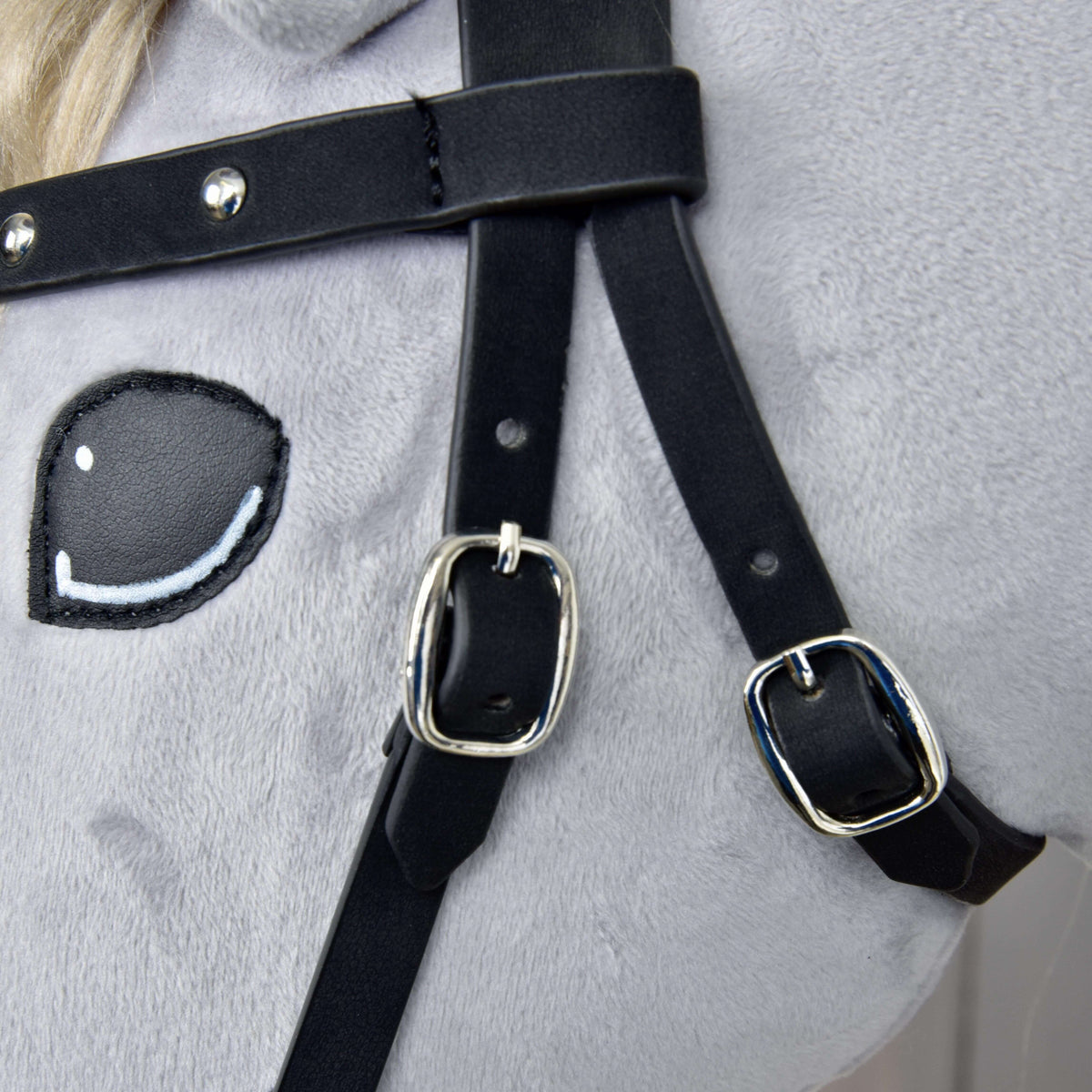 Close-up of the Hobby Horse LarDen Complete Bitless Bridle, Decorated—features soft light gray fabric, an embroidered cartoon eye, and black faux leather straps with shiny silver buckles and detachable reins.
