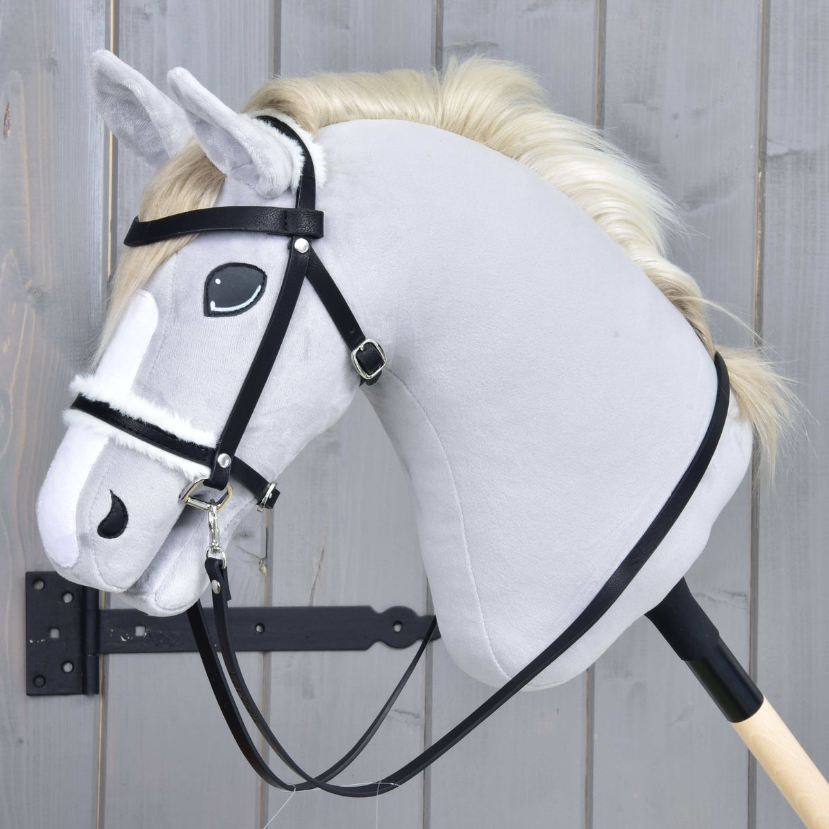 The Hobby Horse LarDen "Complete bridle with fur" features a light gray hobby horse with a cream mane, black leatherette bridle and reins on a wooden stick, displayed against a gray wooden background.