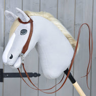 A plush hobby horse from Hobby Horse LarDen features a light gray head, white mane, and a Complete Western bridle One-Ear with split reins, mounted on a wooden stick in front of a gray wooden wall.