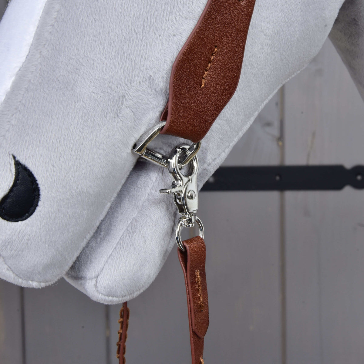 A close-up of the Hobby Horse LarDen Complete Western bridle One-Ear, featuring a brown leather strap with a silver clasp and sliding ear loop, attached to a white plush horse's head on a gray wooden background.
