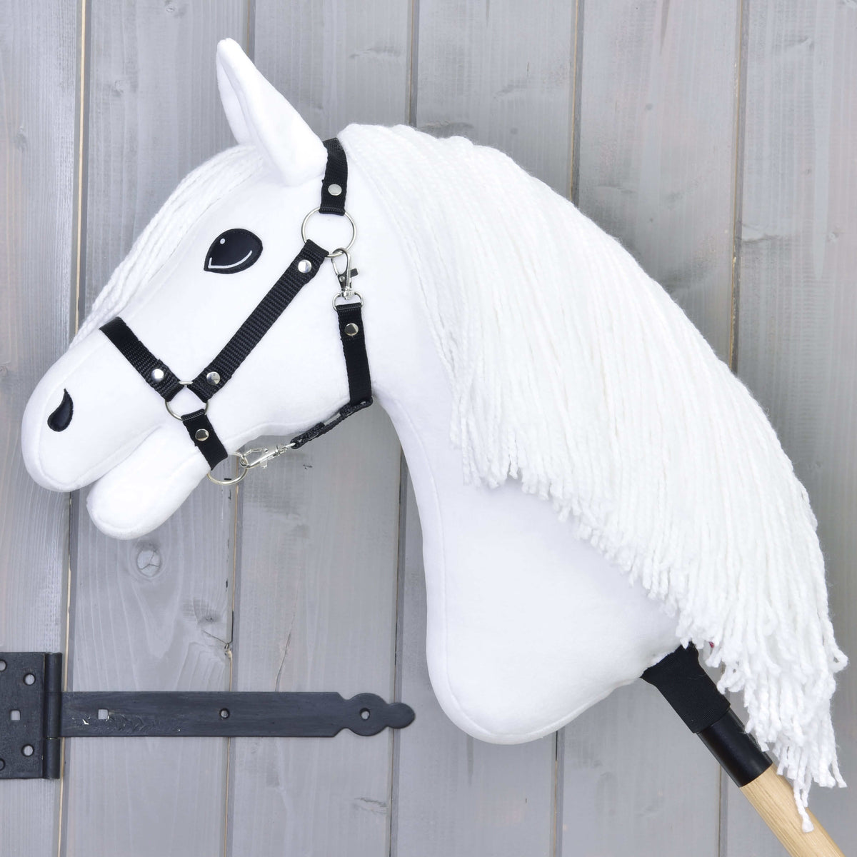The Hobby Horse Lady with Black Halter by Hobby Horse LarDen features a white horse head with a long yarn mane, black halter, and bold black eyes on a wooden stick, displayed against a gray wooden background.