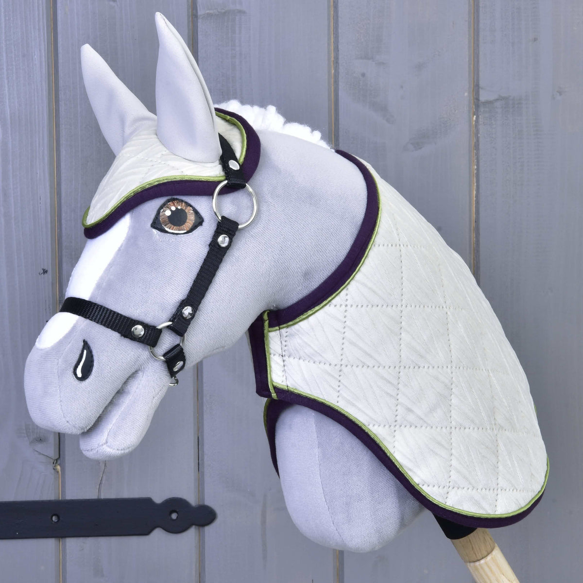 The Set Leonardo by Hobby Horse LarDen features a gray plush hobby horse with a white quilted blanket, matching ear bonnet, black bridle and reins on a wooden stick, shown against a gray wooden wall.