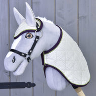 The Set Leonardo by Hobby Horse LarDen features a gray plush hobby horse with a white quilted blanket, matching ear bonnet, black bridle and reins on a wooden stick, shown against a gray wooden wall.