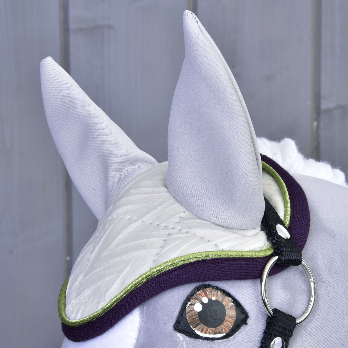 Close-up of the Set Leonardo by Hobby Horse LarDen: a grey fabric horse head with a decorative bridle and white quilted ear bonnet with green edging, displayed against a grey wooden background.