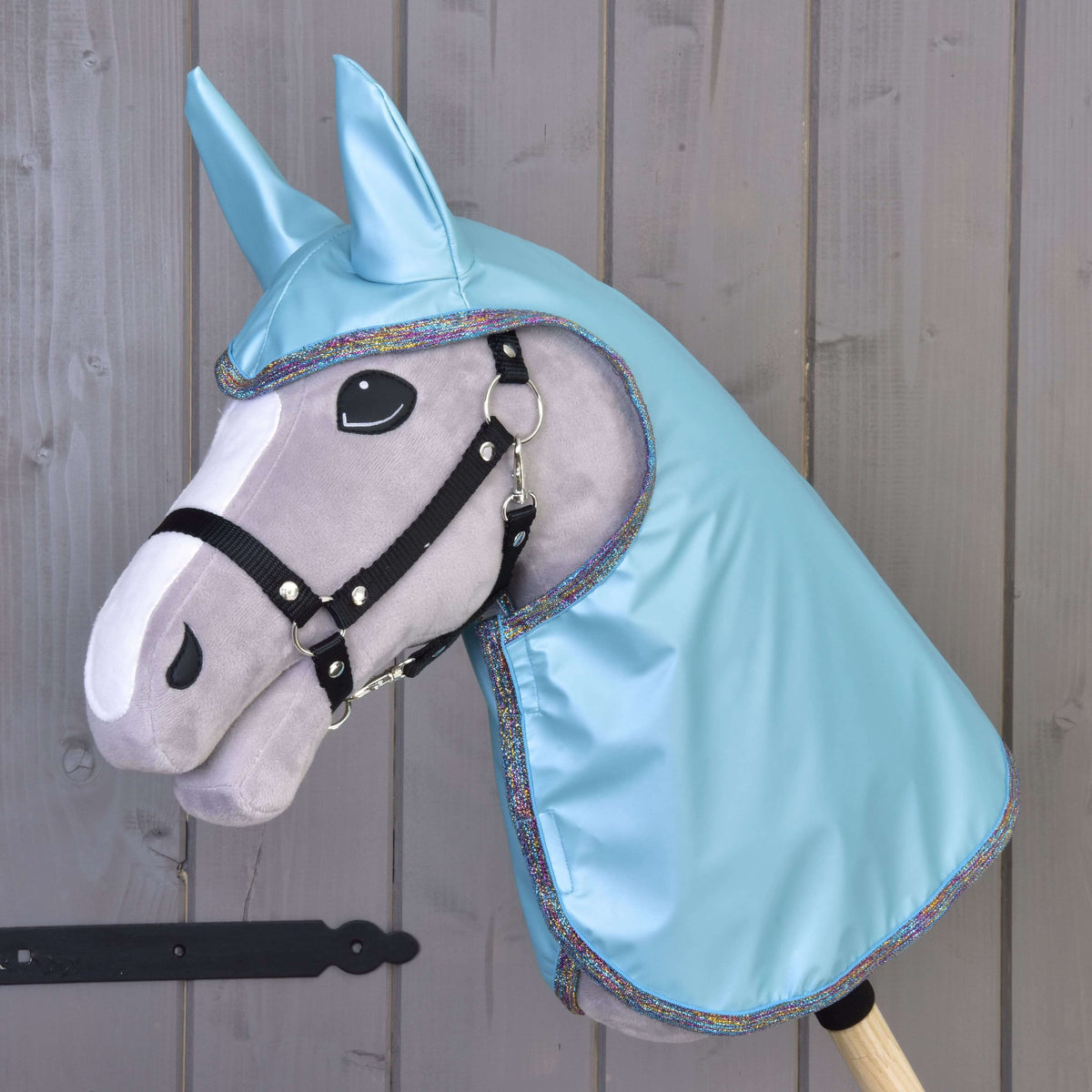 A Hobby Horse LarDen gray hobby horse with a black bridle, large felt eyes, and a Raincoat Light Blue hood with matching ears, displayed against a wooden wall.