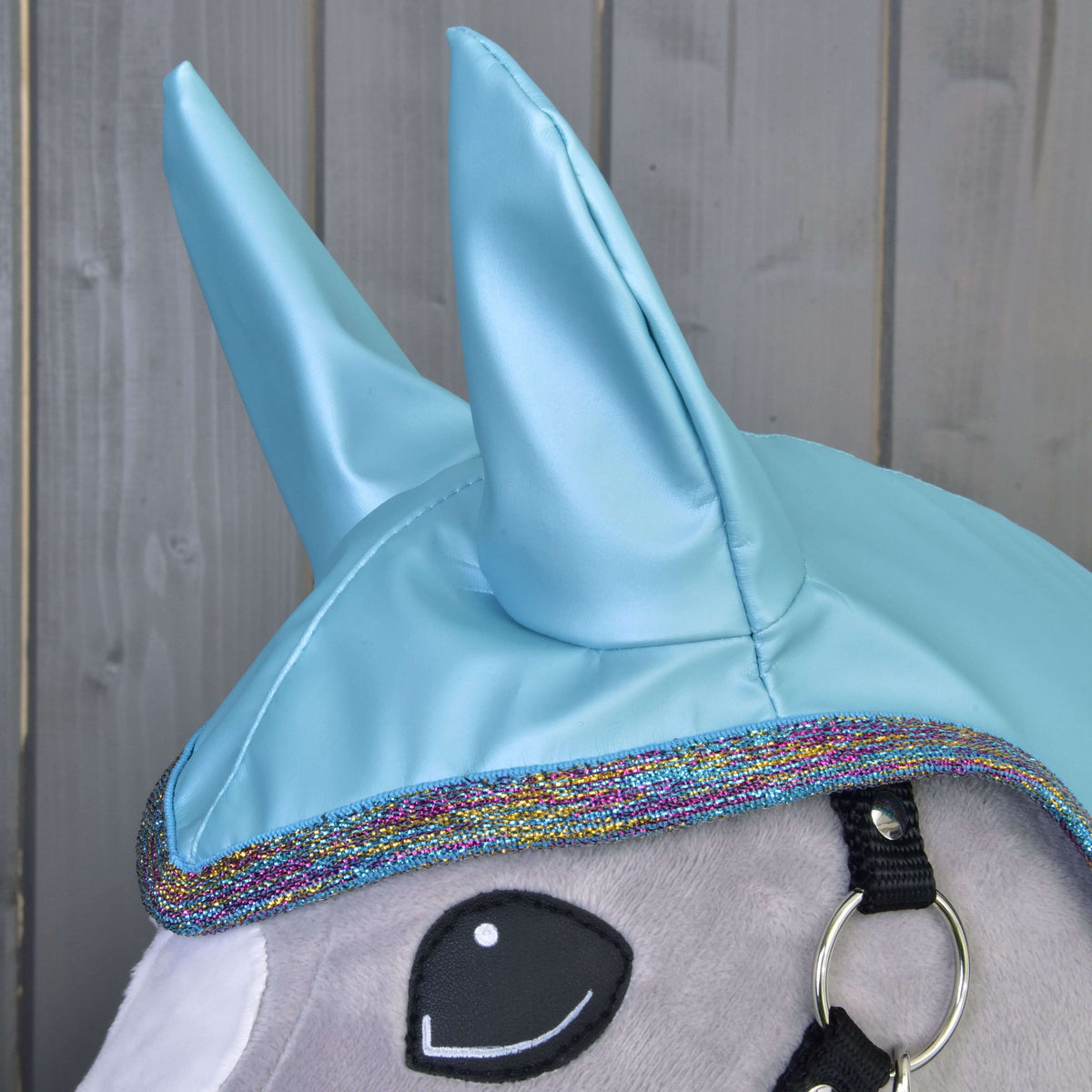 Close-up of the Hobby Horse LarDen Raincoat Light Blue: a plush unicorn head with shiny blue horns, rainbow-trimmed edge, black bridle, and cartoon-style embroidered eye—ideal for all-weather play against wooden paneling.