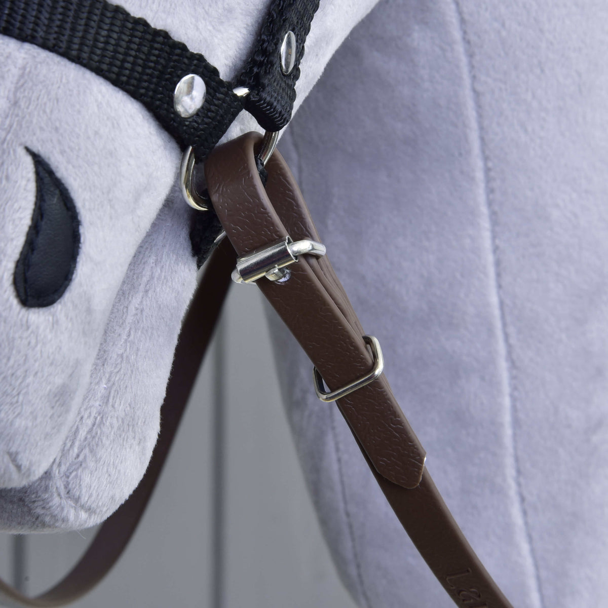 Close-up of Reins Light brown by Hobby Horse LarDen on a grey plush horse toy, showing a leather strap with buckle, black fabric reins and bridle, and a stitched black eye.