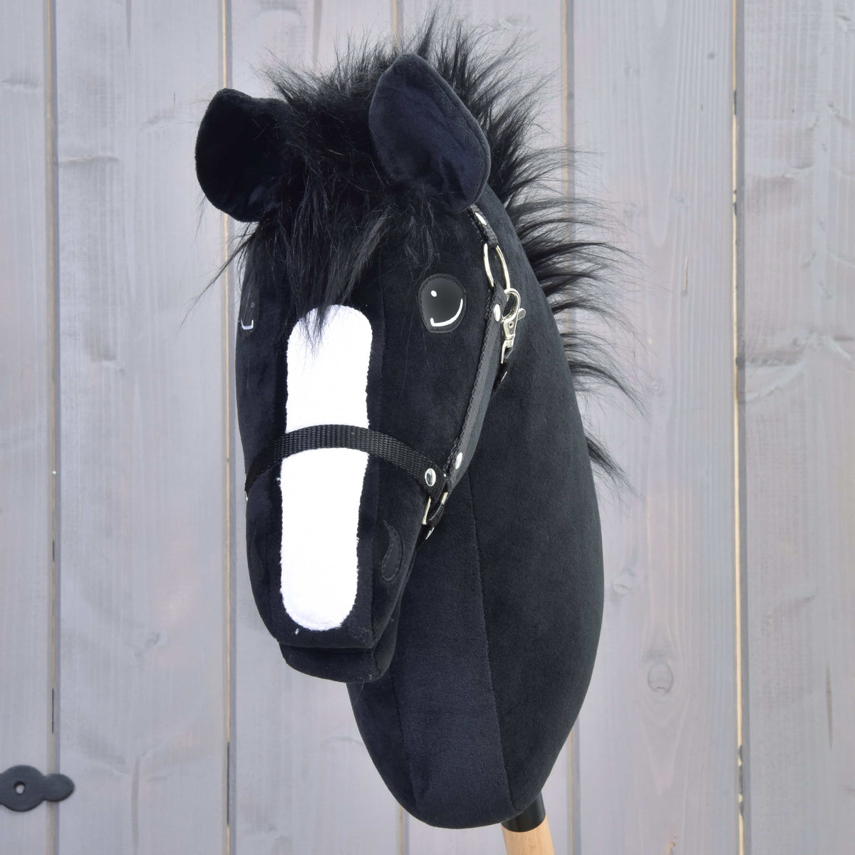 The Hobby Horse LarDen "Hobby Horse Loki with Black Halter" features a black plush Welsh Cob design, white snout, fluffy mane, and wooden stick—perfect for dressage play against a light gray fence backdrop.