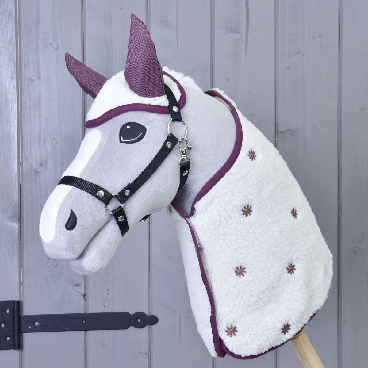 The Hobby Horse LarDen Set Loret features a plush hobby horse with a grey face, black bridle, purple accents, ear bonnet, and a white blanket with small purple flowers, posed against a wooden panel wall.