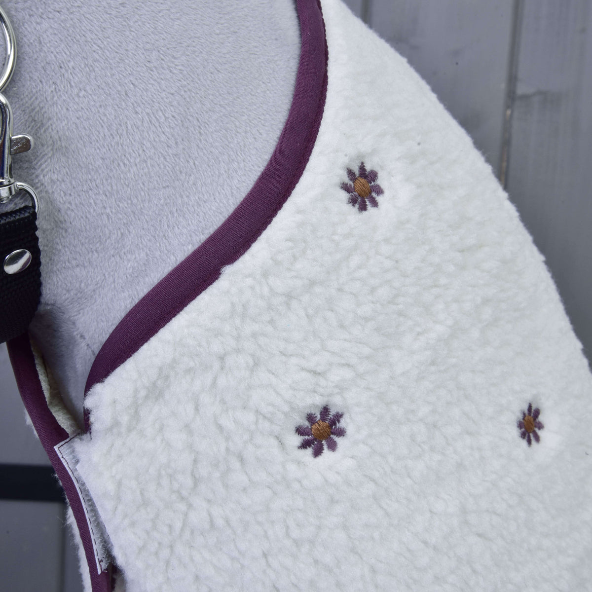 Close-up of Set Loret, a white fleece dog coat by Hobby Horse LarDen, with purple/yellow embroidered flowers and purple trim. Shown on a dog with black leash, it resembles a Hobby Horse blanket, lying on gray wooden surface.