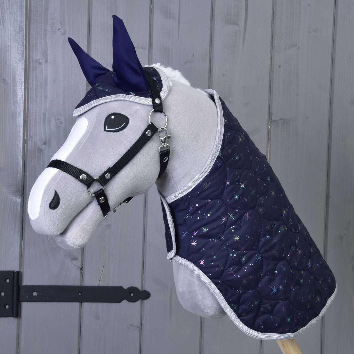The Hobby Horse LarDen Set Magic features a plush stick horse with a navy blue quilted blanket and ear bonnet with star pattern, providing insect protection, shown against a wooden wall.