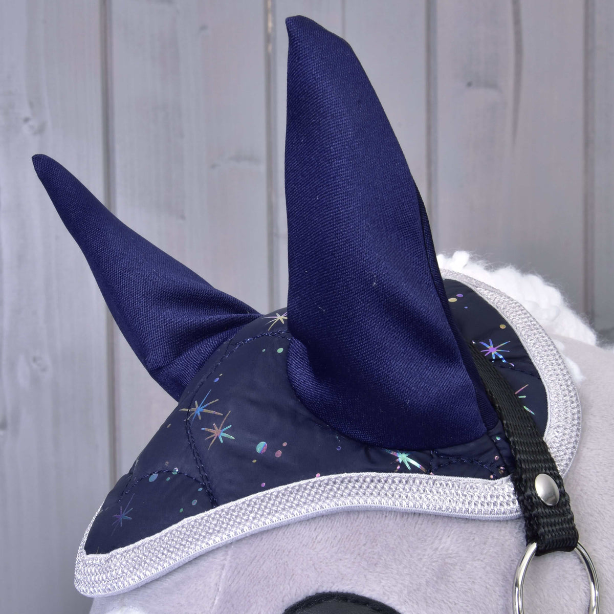 A close-up of the Hobby Horse LarDen Set Magic: a plush horse head with a blue ear bonnet featuring sparkling star patterns and white trim for insect protection, set against a light wooden background.
