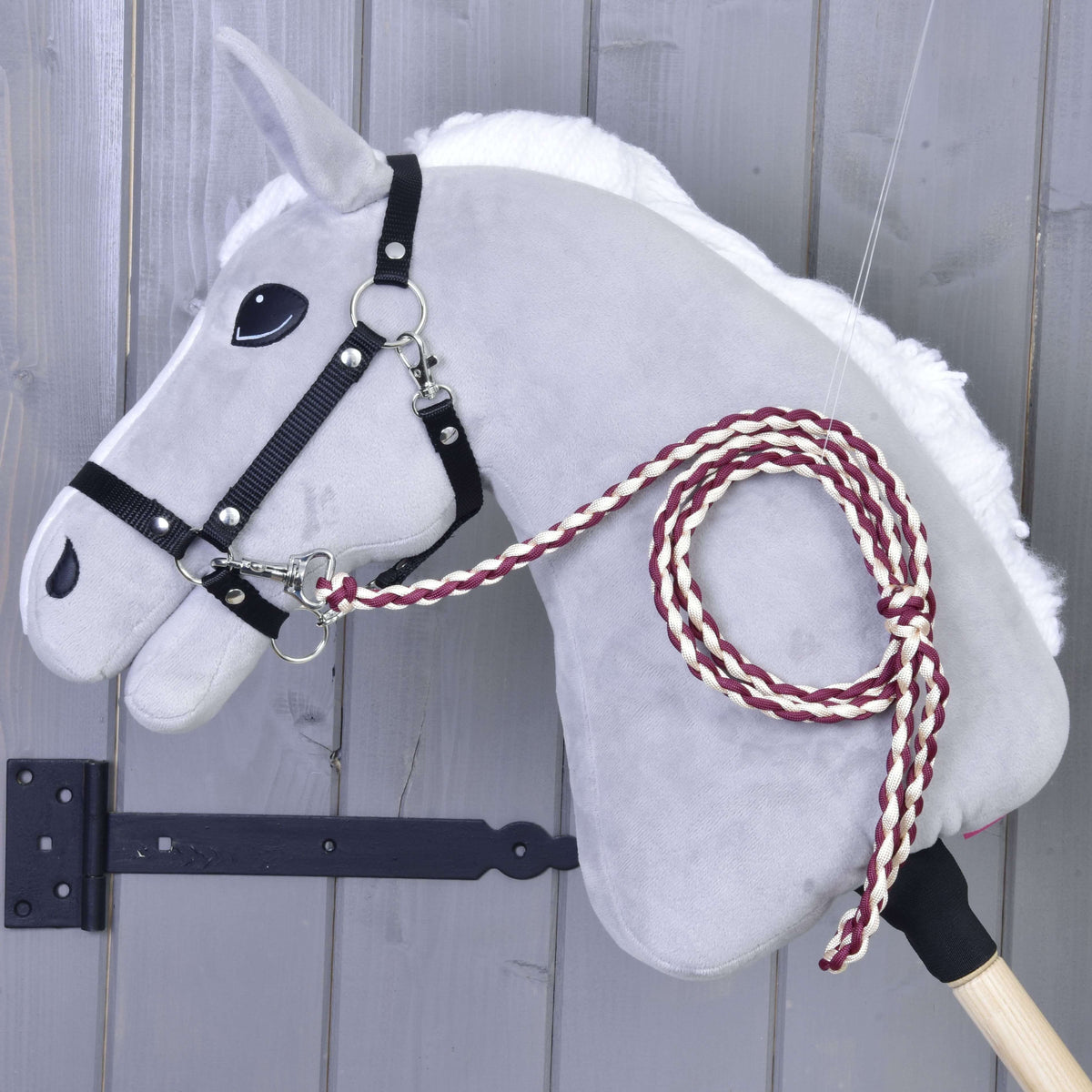 The Hobby Horse LarDen Hobby Horse - Knitted Lead Rope Maroon features a gray felt head with a black bridle and hand-knitted maroon-and-white lead rope, mounted on a wooden stick against a gray wooden wall.