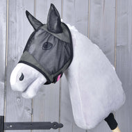 A plush hobby horse with light gray fur wears the Fly Mask De Luxe Grey by Hobby Horse LarDen for extra protection. The background shows a vertical wooden plank wall.