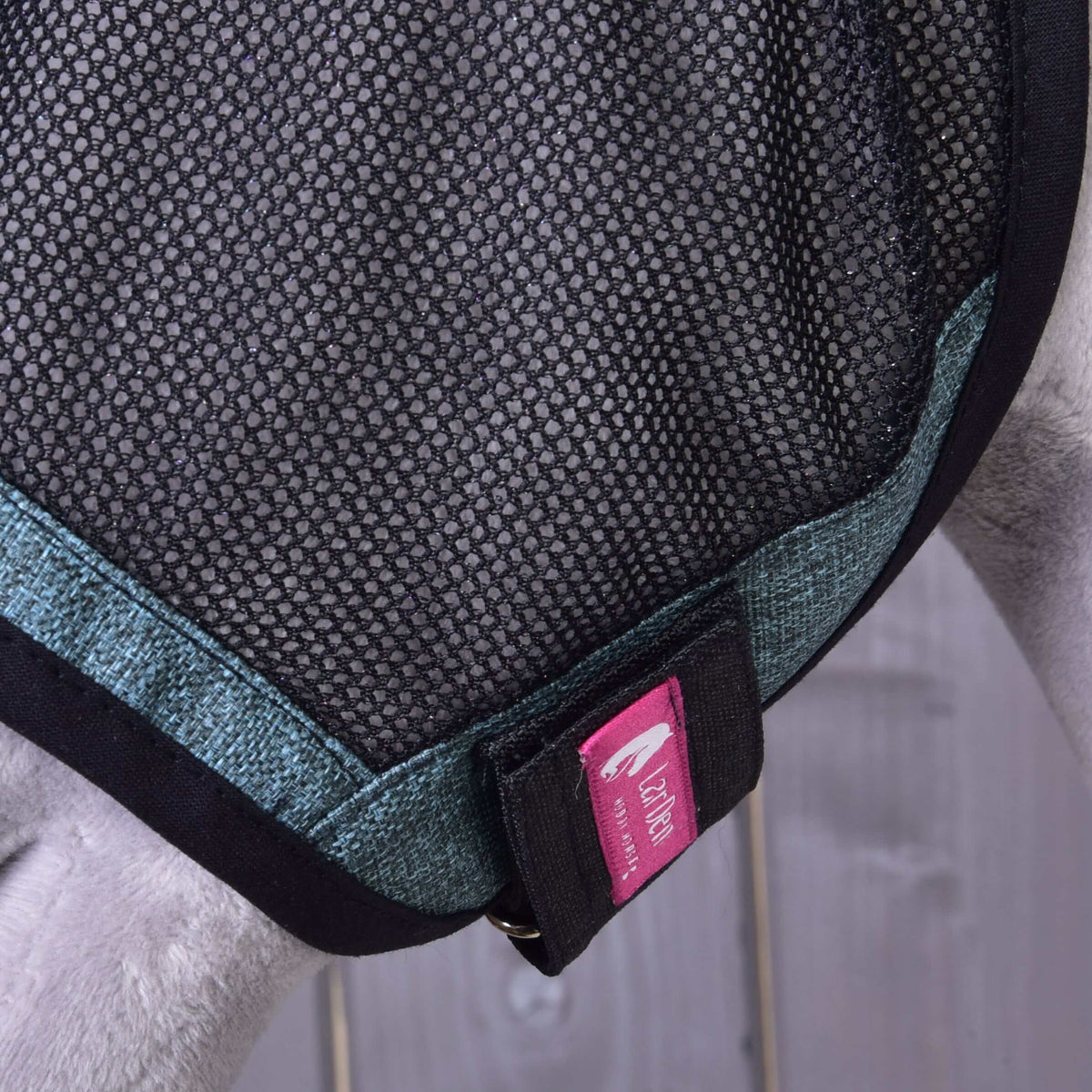 Close-up of a dog in a black mesh harness with blue trim and a pink Zack & Zoey tag. The blurred background resembles the washable material of the Fly Mask De Luxe Turquoise by Hobby Horse LarDen.