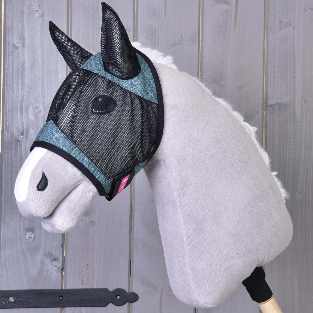 The Hobby Horse LarDen Fly Mask De Luxe Turquoise is displayed on a gray plush hobby horse with a white muzzle, shown posed against wooden planks. The washable mask offers stylish protection for your hobby horse.