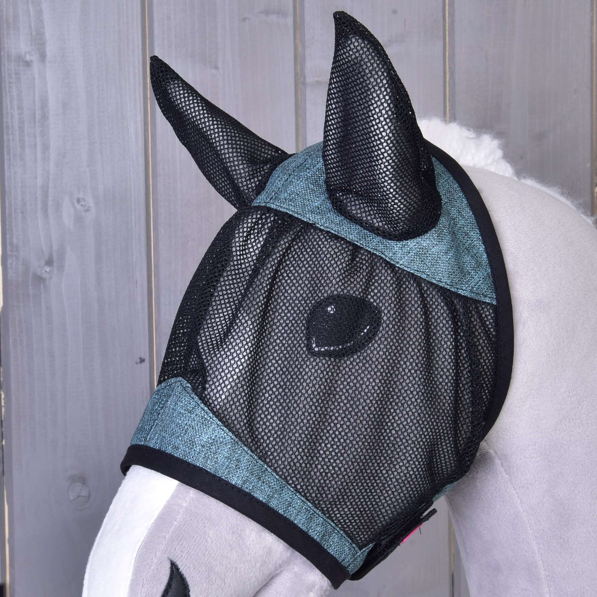 A close-up of a horse wearing the Fly Mask De Luxe Turquoise by Hobby Horse LarDen, a black mesh, washable fly mask with teal trim that covers its eyes and ears for protection. Wooden paneling is in the background.