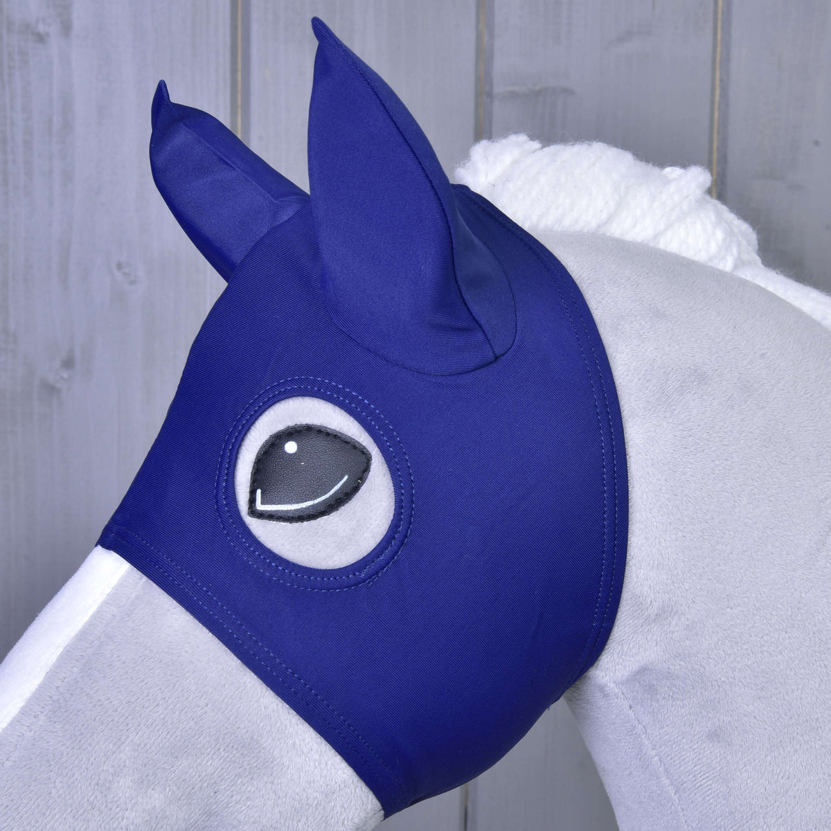 A close-up of a gray horse wearing the Horse Mask LarDen by Hobby Horse LarDen, featuring ear covers and eye cutouts, standing in front of a wooden background.