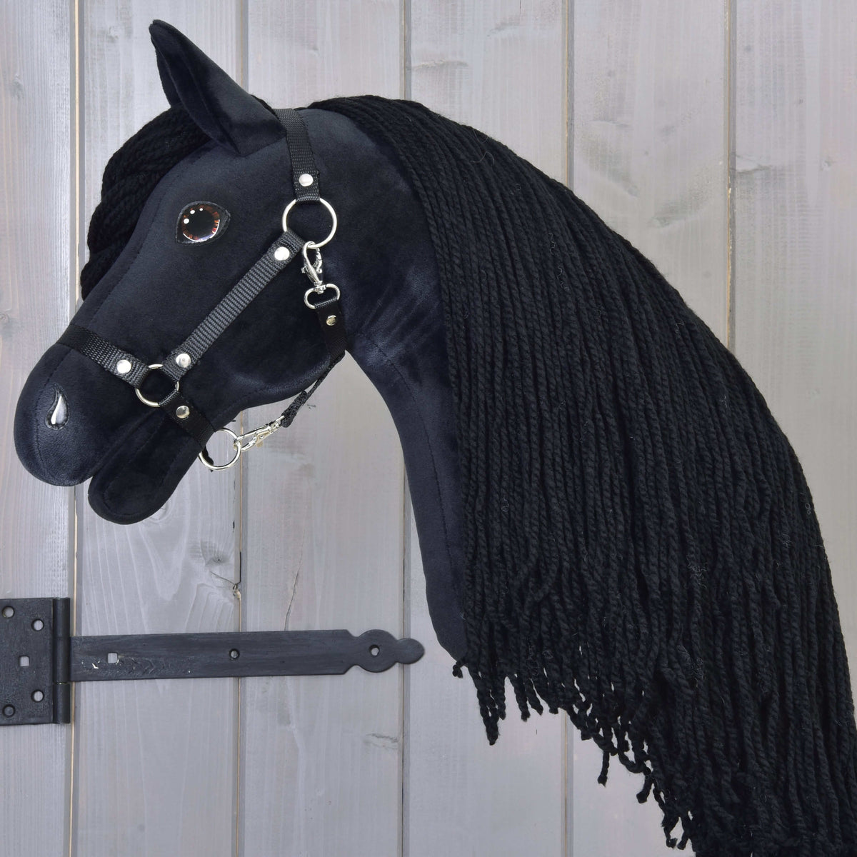 The Hobby Horse Maximus with Black Halter by Hobby Horse LarDen, featuring a plush black Friesian design, long braided mane, bridle with metal rings, and shiny button eye, hangs on a wooden wall.