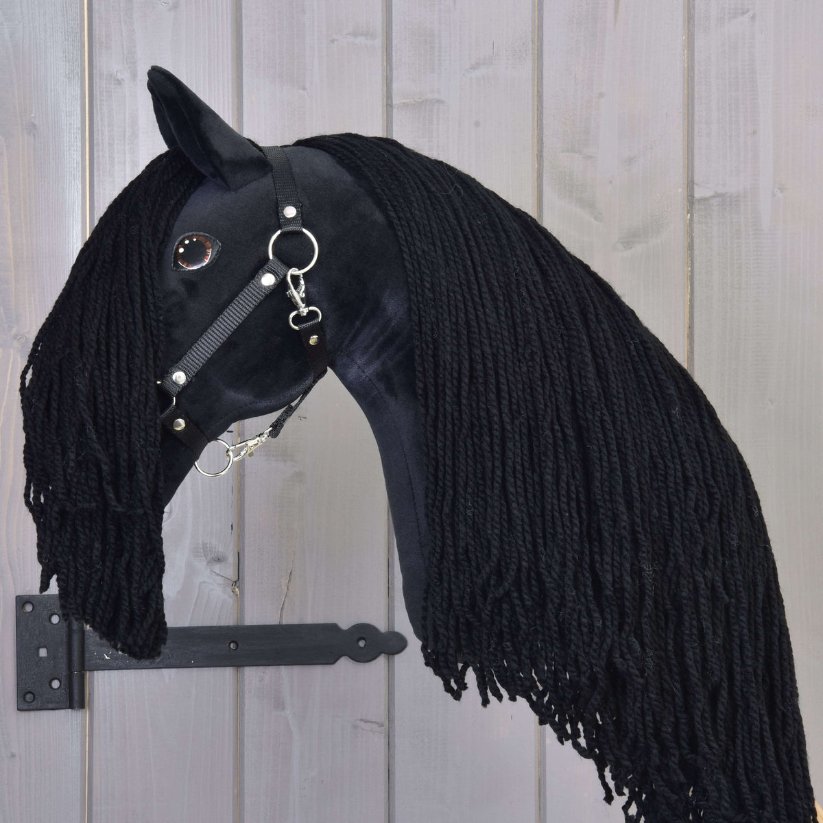 The Hobby Horse Maximus with Black Halter by Hobby Horse LarDen is a handmade black stallion toy featuring a long yarn mane, shiny black bridle, and button eye, mounted on wood with a black metal bracket.